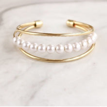 Load image into Gallery viewer, Elaine Pearl Bracelet features three thin, smooth bands, with a row of pearls in the middle. Beautiful and simple, this bracelet is the perfect feminine complement.  14k Gold plated on Brass, Synthetic Pearls Diameter: 2.5 inches
