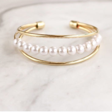 Elaine Pearl Bracelet features three thin, smooth bands, with a row of pearls in the middle. Beautiful and simple, this bracelet is the perfect feminine complement.  14k Gold plated on Brass, Synthetic Pearls Diameter: 2.5 inches